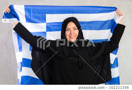 Balanced smiling Muslim woman in traditional black hijab holds flag of Greece 102211356