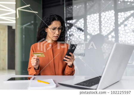 Upset and disappointed hispanic businesswoman inside office at workplace, woman holding bank credit card and phone, employee cheated trying to make online purchase, money transfer error. Upset and disappointed hispanic businesswoman inside office at workplace, woman holding bank credit card and phone, employee cheated trying to make online purchase, money transfer error. 102225597