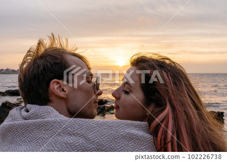 Portraits of lovers, romantic couple of lovers hugging, kissing, touching, eye contact at sunset, sunrise against the background of the sea, sun, clouds in fiery red, orange colors Portraits of lovers, romantic couple of lovers hugging, kissing, touching, eye contact at sunset, sunrise against the background of the sea, sun, clouds in fiery red, orange colors 102226738