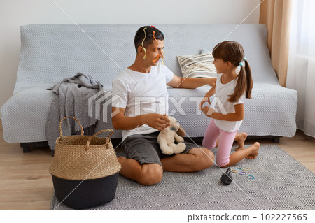 Portrait of dark haired male with colored tresses with his female child playing together at home, sitting on floor near cough in light room, happy time during weekend. Portrait of dark haired male with colored tresses with his female child playing together at home, sitting on floor near cough in light room, happy time during weekend. 102227565