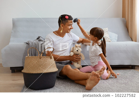 Image of young adult father spending time with his daughter while sitting on floor near sofa in living room, kid making hairstyle for dad with colored tresses, combing hair. 102227582