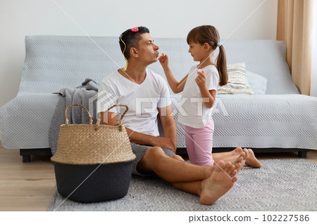 Portrait of positive happy man wearing white casual style T-shirt and jeans short sitting on floor with child, daughter making makeup with lipstick, having fun together. 102227586