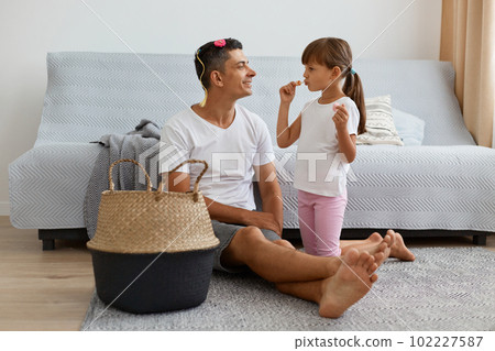 Indoor shot of attractive Caucasian man wearing white casual style T-shirt and jeans short sitting on floor with little dark haired girl, daughter using mom's pomade, spending time with dad. Indoor shot of attractive Caucasian man wearing white casual style T-shirt and jeans short sitting on floor with little dark haired girl, daughter using mom's pomade, spending time with dad. 102227587