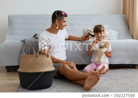 Image of Caucasian man wearing white casual style t shirt sitting on floor near sofa with child, father talking with his daughter after playing together as hairdressers. 102227588