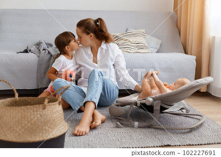 Full length portrait of happy female with dark hair and ponytail, wearing casual clothing sitting on floor with her kids, kissing her charming daughter, taking care of infant baby in rocking chair. 102227691