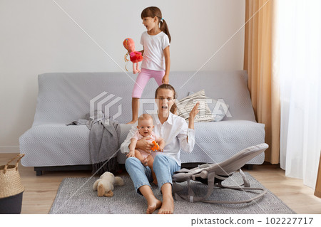 Portrait of exhausted Caucasian woman sitting on floor near sofa and screaming why, being tired of noisy little children, wearing white t shirt and jeans, posing in living room. 102227717