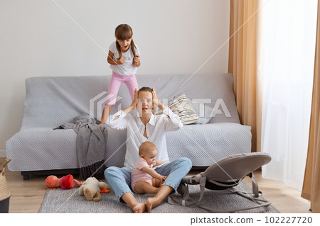 Indoor shot of tired young adult man sitting on floor near sofa and covering ears with palms, being tired of noisy screaming children, needs time to rest. Indoor shot of tired young adult man sitting on floor near sofa and covering ears with palms, being tired of noisy screaming children, needs time to rest. 102227720