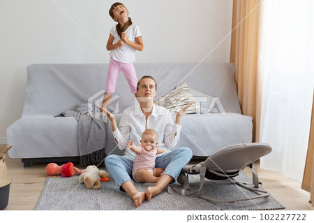 Full length of portrait of depressed mother wearing white shirt and jeans, sitting on floor near sofa and feels desperate, raised arm, don't know what to with screaming children. 102227722