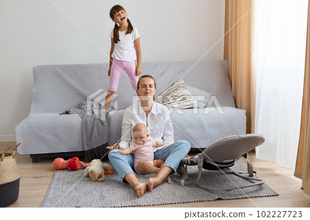 Full length portrait of sad desperate woman wearing white shirt and jeans sitting on floor with her daughters, being depressed and tired looking after noisy kids. Full length portrait of sad desperate woman wearing white shirt and jeans sitting on floor with her daughters, being depressed and tired looking after noisy kids. 102227723
