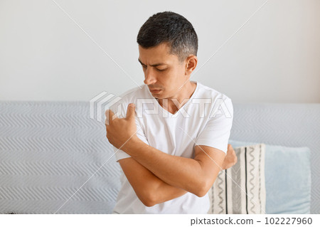 Indoor shot of ill sick man wearing white T-shirt sitting on sofa in living room, holding painful shoulder, gets trauma, suffering hand sprain or arthritis inflammation. 102227960