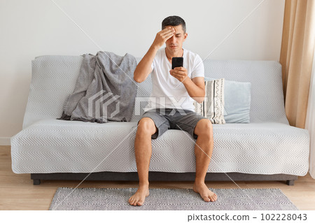 Image of Caucasian brunette man wearing white t shirt and jeans shirt sitting on sofa at home, holding smart phone, reading news in social networks, keeping hand on his head. Image of Caucasian brunette man wearing white t shirt and jeans shirt sitting on sofa at home, holding smart phone, reading news in social networks, keeping hand on his head. 102228043