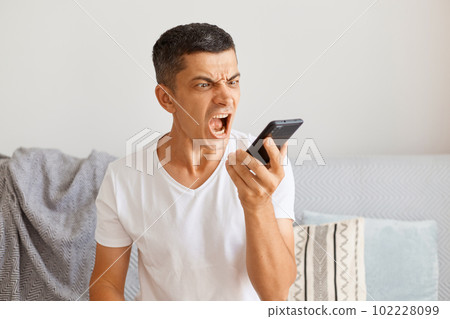 Horizontal shot of dark haired young adult male wearing white casual style t shirt sitting on sofa, holding cell phone in hands and screaming with anger. 102228099