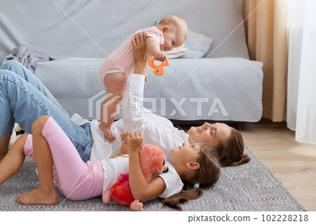 Side view portrait of mom and sweet child girls lying and playing on floor, mother holding infant baby ina hands, family expressing positive emotions and happiness. 102228218