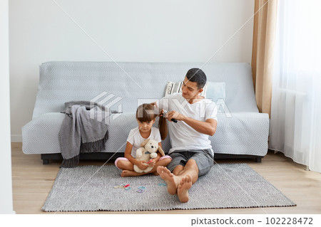 Portrait of young adult Caucasian man posing at home with his cute daughter, making pigtails or ponytail for little kid, family sitting on floor near sofa and window. Portrait of young adult Caucasian man posing at home with his cute daughter, making pigtails or ponytail for little kid, family sitting on floor near sofa and window. 102228472