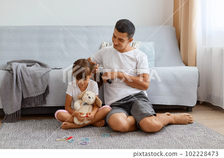 Indoor shot man with dark hair wearing white t shirt combing daughter's hair while sitting on floor near cough, preparing for going to school, expressing positive emotions, kid holding toy. 102228473