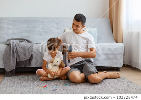 Image of caring father wearing white t shirt sitting on floor and making hairstyle for his cute little daughter, looking at child with pleasant smile, combing hair, makes ponytail. 102228474