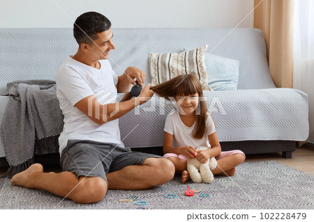 Indoor shot of attractive man wearing white t shirt sitting on floor and making hairstyle for his cute little daughter, taking care of his female child, spending pleasant time together. 102228479