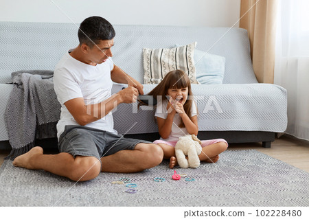 Indoor shot of attractive man wearing white casual style T-shirt and jeans short sitting on floor with his daughter, father making hairstyle for charming kid, parenthood. Indoor shot of attractive man wearing white casual style T-shirt and jeans short sitting on floor with his daughter, father making hairstyle for charming kid, parenthood. 102228480