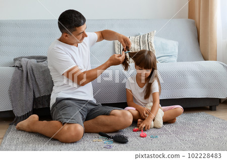Indoor shot of young adult attractive man wearing white t shirt and jeans sitting on floor near sofa and making hairstyle for his cute daughter, preparing for going to school. Indoor shot of young adult attractive man wearing white t shirt and jeans sitting on floor near sofa and making hairstyle for his cute daughter, preparing for going to school. 102228483