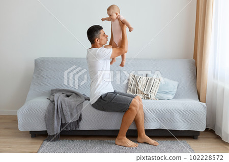 Indoor shot of ghandsome brunette father holding infant baby girl in hands and understand they need changing baby's diaper, feeling bad smell, raising kid up air. Indoor shot of ghandsome brunette father holding infant baby girl in hands and understand they need changing baby's diaper, feeling bad smell, raising kid up air. 102228572