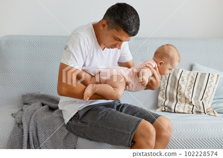 Portrait of attractive man father sitting on sofa and holding his infant daughter, smelling, understand that is time to change diaper, male wearing white t shirt and jeans short. Portrait of attractive man father sitting on sofa and holding his infant daughter, smelling, understand that is time to change diaper, male wearing white t shirt and jeans short. 102228574