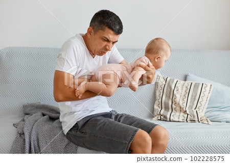 Portrait of frustrated man posing with toddler girl in hands, smelling baby, needs to change diaper and wash kid, sitting on sofa in light living room at home. Portrait of frustrated man posing with toddler girl in hands, smelling baby, needs to change diaper and wash kid, sitting on sofa in light living room at home. 102228575