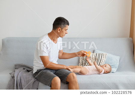 Indoor shot of handsome brunette father playing with his infant daughter while sitting on sofa, wearing white t shirt and jeans short, looking at baby with serious facial expression. Indoor shot of handsome brunette father playing with his infant daughter while sitting on sofa, wearing white t shirt and jeans short, looking at baby with serious facial expression. 102228576