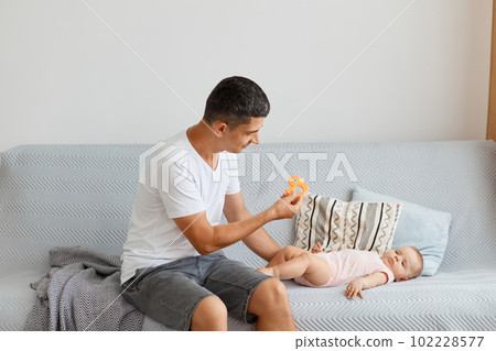 Good looking Caucasian attractive young adult man sitting sofa and playing with his little kid, looking at baby with smile and great love, wearing white t shirt and jeans short. Good looking Caucasian attractive young adult man sitting sofa and playing with his little kid, looking at baby with smile and great love, wearing white t shirt and jeans short. 102228577