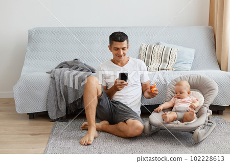 Indoor shot of smiling man wearing white casual style T-shirt and jeans short sitting on floor with daughter in rocking chair, father using smart phone and showing toy for kid. Indoor shot of smiling man wearing white casual style T-shirt and jeans short sitting on floor with daughter in rocking chair, father using smart phone and showing toy for kid. 102228613