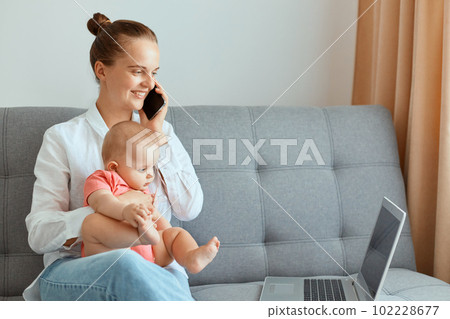Indoor shot of positive optimistic woman with hair bun wearing white shirt and jeans sitting on sofa with infant baby daughter, having pleasant conversation with friend while working on notebook. Indoor shot of positive optimistic woman with hair bun wearing white shirt and jeans sitting on sofa with infant baby daughter, having pleasant conversation with friend while working on notebook. 102228677