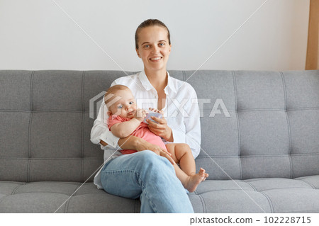 Image of smiling happy woman with bun hairstyle wearing white shirt and jeans, posing with her small daughter, looking at camera with positive facial expression. 102228715