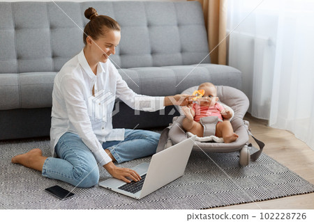 Indoor shot of smiling satisfied woman with bun hairstyle wearing white shirt and jeans, posing at home while sitting on floor, working on laptop and showing toy to infant kid in rocking chair. 102228726