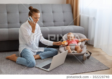 Indoor shot of attractive woman in white t shirt sitting near cough with baby daughter in rocking chair and working on laptop, talking on phone with business partner. Indoor shot of attractive woman in white t shirt sitting near cough with baby daughter in rocking chair and working on laptop, talking on phone with business partner. 102228740