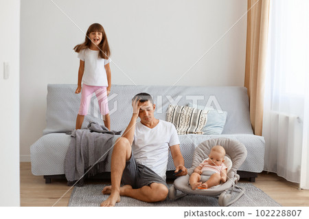 Indoor shot of tired attractive young adult man sitting on floor near sofa, having headache, exhausted of loud kids' playing, wearing white t shirt and jeans short. Indoor shot of tired attractive young adult man sitting on floor near sofa, having headache, exhausted of loud kids' playing, wearing white t shirt and jeans short. 102228807