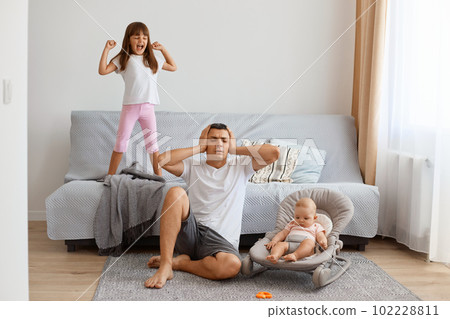 Exhausted handsome brunette father sitting on floor near sofa with smaller daughter in bouncer, wearing white t shirt and jeans short, man covering ears from screaming elder daughter. Exhausted handsome brunette father sitting on floor near sofa with smaller daughter in bouncer, wearing white t shirt and jeans short, man covering ears from screaming elder daughter. 102228811