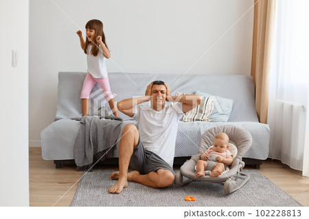 Indoor shot of young adult man sitting on floor near sofa and covering ears with palms, being tired of loud daughter jumping on floor and scream, wearing white t shirt and jeans short. Indoor shot of young adult man sitting on floor near sofa and covering ears with palms, being tired of loud daughter jumping on floor and scream, wearing white t shirt and jeans short. 102228813
