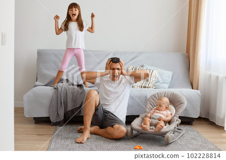 Tired screaming handsome brunette father spending time with daughters while sitting on floor near sofa, covering ears with hands, wants not to hear the screaming older daughter. Tired screaming handsome brunette father spending time with daughters while sitting on floor near sofa, covering ears with hands, wants not to hear the screaming older daughter. 102228814