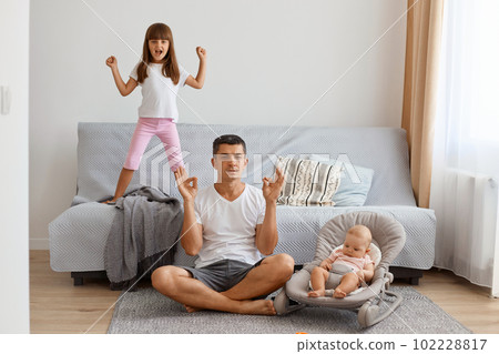 Horizontal shot of sdepressed attractive young adult man sitting on floor in lotus pose near sofa and looking after his little kids, practising yoga, trying to relax. Horizontal shot of sdepressed attractive young adult man sitting on floor in lotus pose near sofa and looking after his little kids, practising yoga, trying to relax. 102228817
