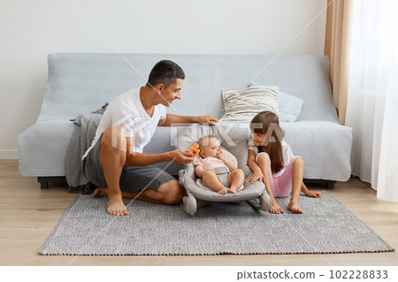 Indoor shot of happy smiling man wearing white casual style T-shirt and jeans short sitting on floor with his two daughters, newborn baby in rocking chair and elder kid with dark hair. Indoor shot of happy smiling man wearing white casual style T-shirt and jeans short sitting on floor with his two daughters, newborn baby in rocking chair and elder kid with dark hair. 102228833