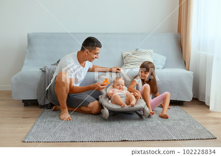 Indoor shot of brunette young father with his daughters sitting on floor near gray sofa, man with elder kid trying to calm down crying little infant baby in rocking chair. Indoor shot of brunette young father with his daughters sitting on floor near gray sofa, man with elder kid trying to calm down crying little infant baby in rocking chair. 102228834