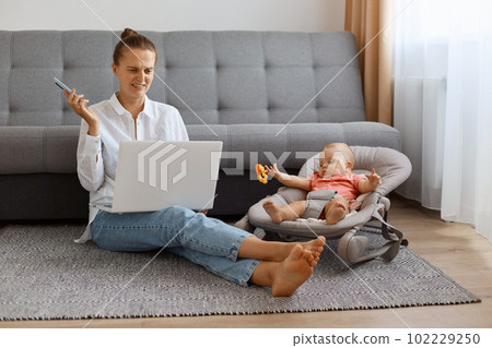 Indoor shot of young adult female wearing white shirt looking at computer screen with disguast, frowning face, spending time with her infant kid in bouncer. Indoor shot of young adult female wearing white shirt looking at computer screen with disguast, frowning face, spending time with her infant kid in bouncer. 102229250