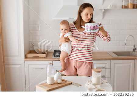 Portrait of young adult woman standing at the table in the kitchen with baby daughter in hands, being ready to cook or bake, holding pot and smelling what inside. 102229256