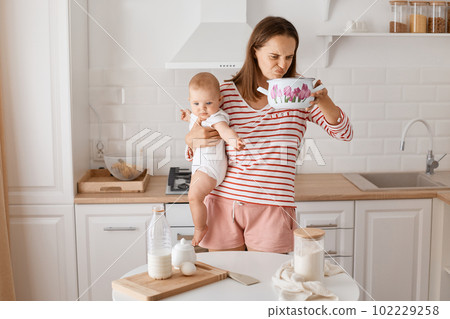 Indoor shot of attractive dark haired female wearing striped shirt and short posing in kitchen with her infant daughter, holding pot and smelling, feeling bed odor. 102229258