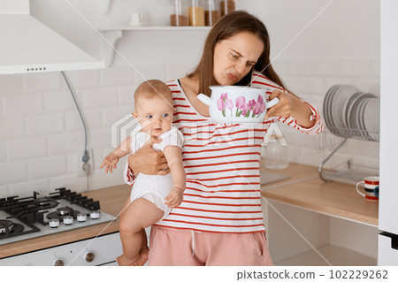 Young adult Caucasian woman standing at table in the kitchen and smelling food in pot while talking on mobile phone and holding infant baby daughter in hands. 102229262