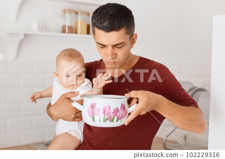 Image of brunette Caucasian man wearing maroon casual style t shirt standing in kitchen with baby daughter in hands, smelling soup in pot, feels awful odor, needs cook fresh food. 102229316