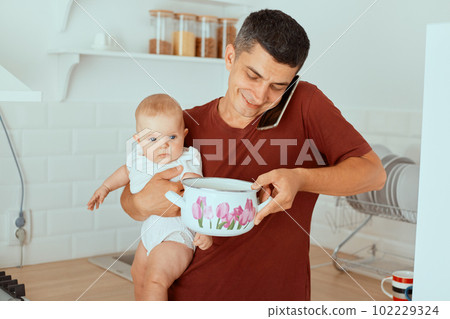 Indoor shot of smiling dark haired father talking via mobile phone and holding baby daughter and pot in hands, man taking care of his kid, being in paternity leave. Indoor shot of smiling dark haired father talking via mobile phone and holding baby daughter and pot in hands, man taking care of his kid, being in paternity leave. 102229324