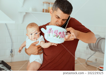 Indoor shot of handsome father with infant baby in hands, standing in kitchen and holding pot with expired food, smelling, feeling bad odor, talking phone with wife, asking what to cook. Indoor shot of handsome father with infant baby in hands, standing in kitchen and holding pot with expired food, smelling, feeling bad odor, talking phone with wife, asking what to cook. 102229325