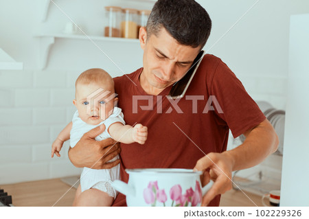 Horizontal shot of dark haired Caucasian young adult man wearing casual style t shirt holding baby girl and pot with soup or porridge, talking phone, asking wife how to feed daughter. Horizontal shot of dark haired Caucasian young adult man wearing casual style t shirt holding baby girl and pot with soup or porridge, talking phone, asking wife how to feed daughter. 102229326