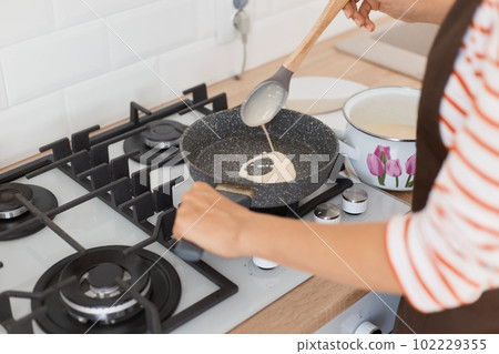 Portrait of of woman's hand fry pancake on frying pan, holding spoon, making breakfast for her family, unknown female wearing striped shier and apron. Portrait of of woman's hand fry pancake on frying pan, holding spoon, making breakfast for her family, unknown female wearing striped shier and apron. 102229355
