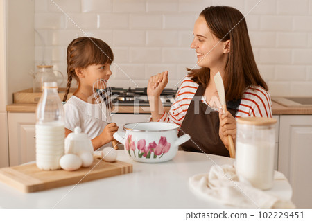Indoor shot of happy satisfied young adult woman cooking with her daughter in the kitchen, having fun together with her charming kid, expressing positive emotions. 102229451
